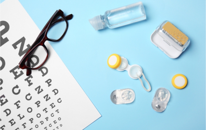 A contact lens care kit laid next to a pair of glasses and eye chart on a blue background.