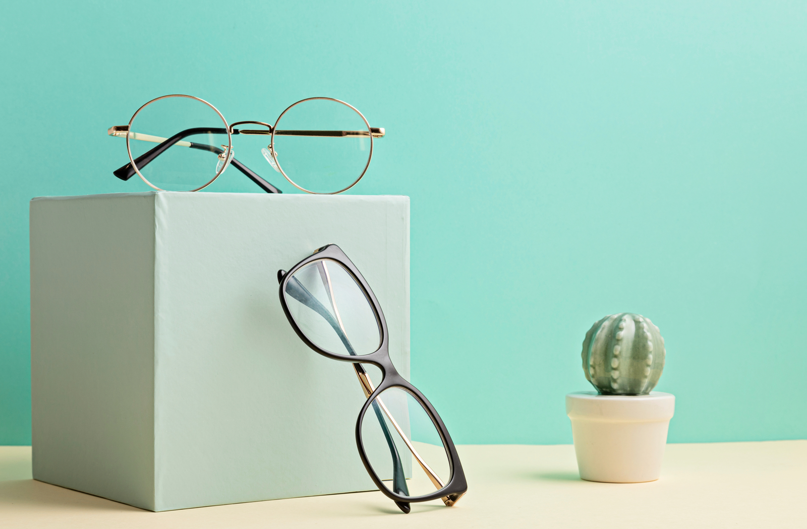 Two pairs of glasses lay on a light green box next to a light green background.