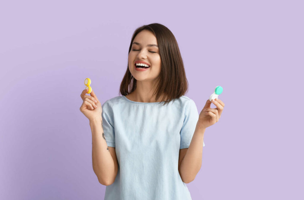 Happy young woman standing against a purple background and holding a contact lens case in each hand.