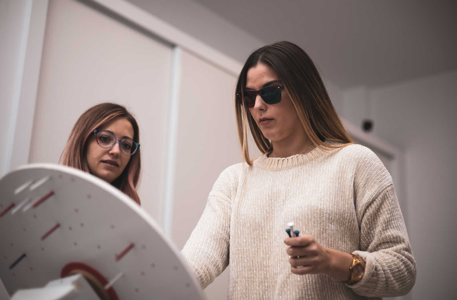 An adult woman is wearing 3D glasses while undergoing vision therapy and is supervised by a female optician on her side.
