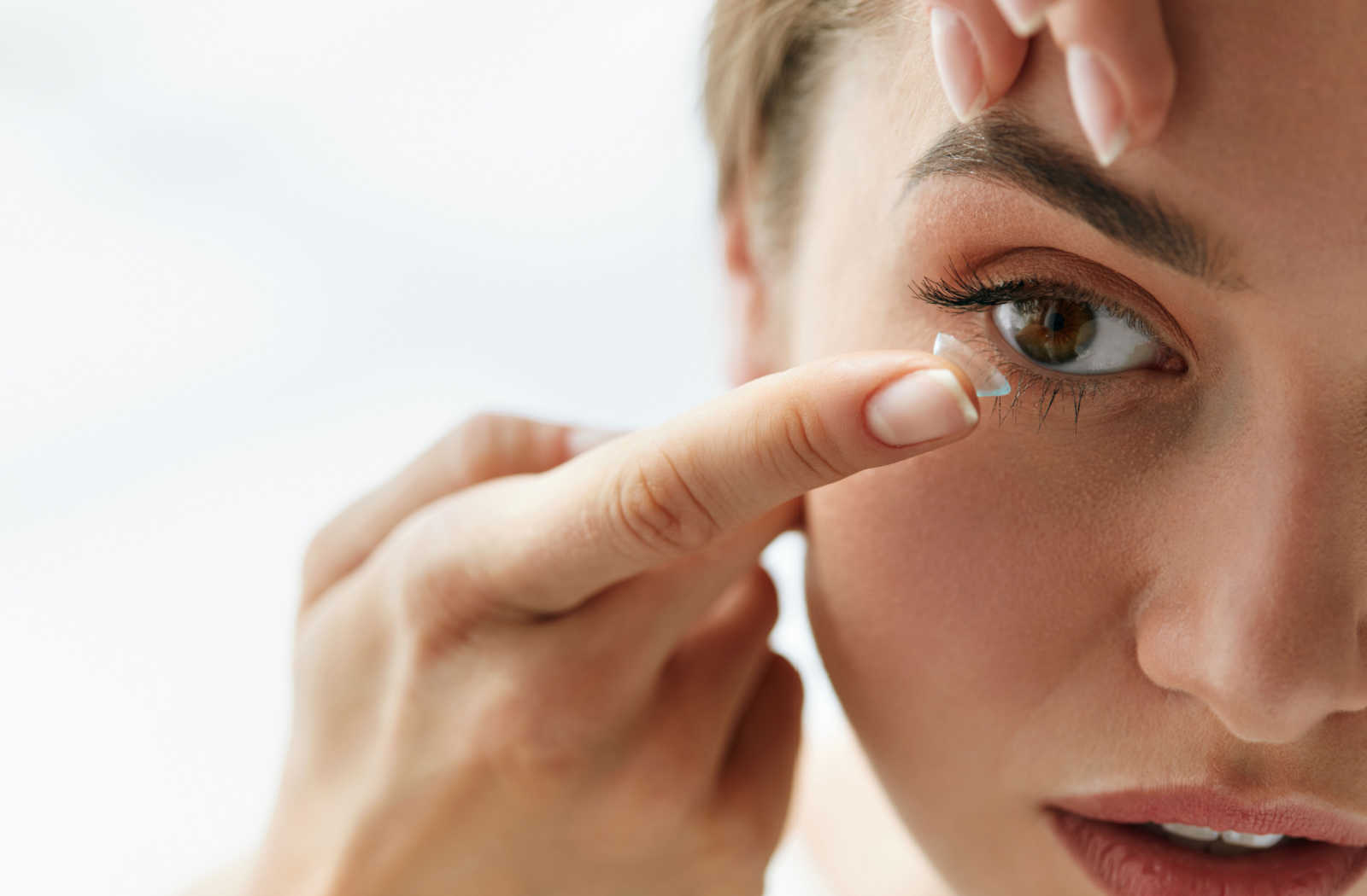 A woman putting a contact lens on her right eye using her right hand with her left hand holding her eyelid.