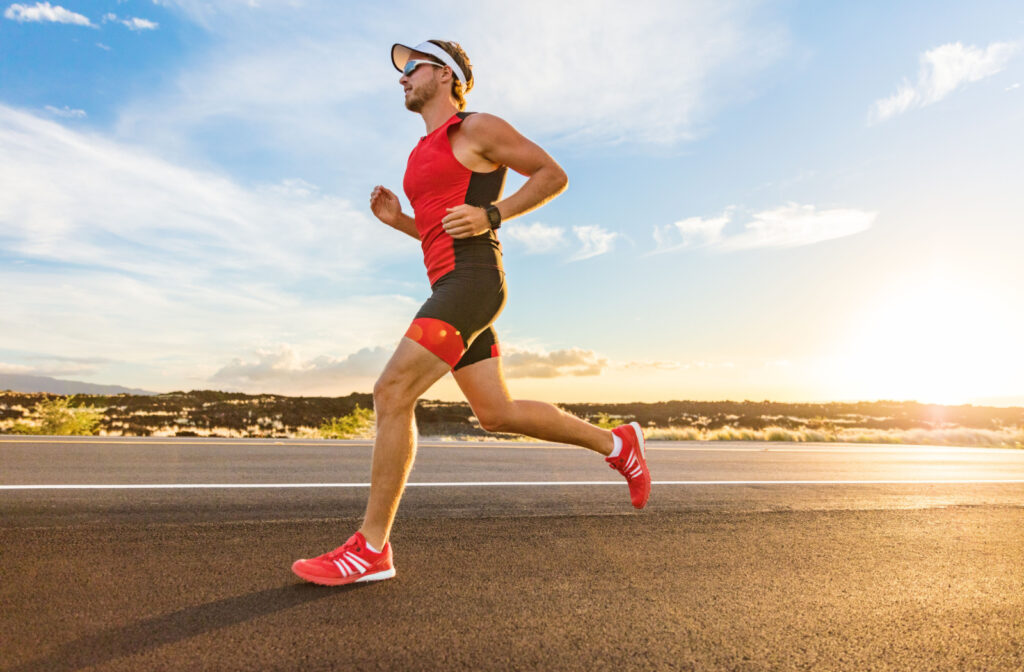 A fit man running on the side of a road, wearing red and black spandex, red shoes, a visor, and sunglasses.