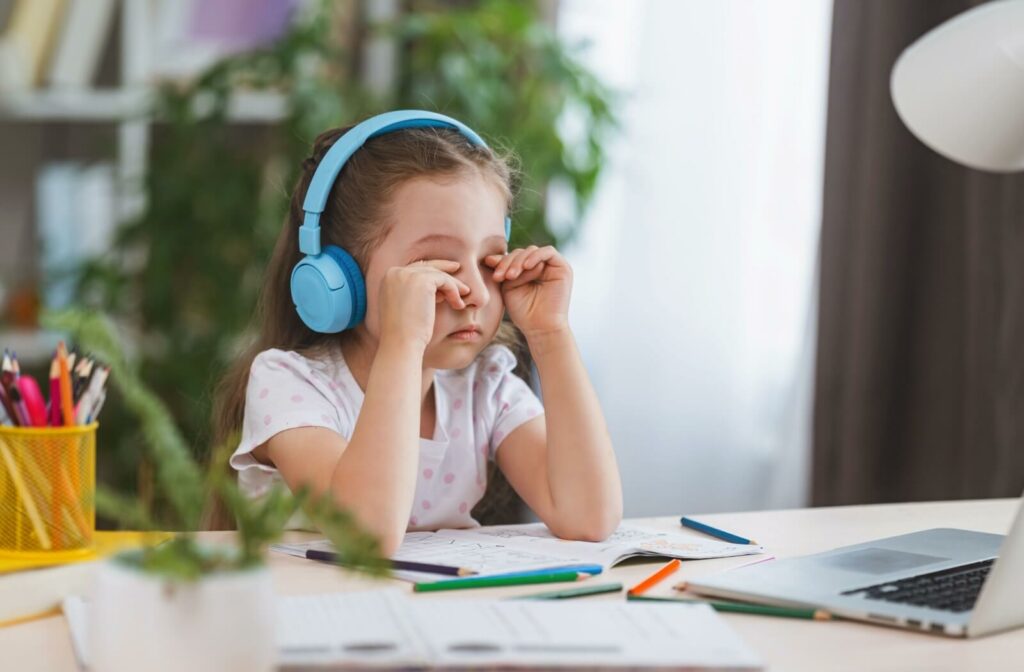 A child with headphones on rubbing their eyes from convergence insufficiency while doing homework on a desk in front of a laptop.