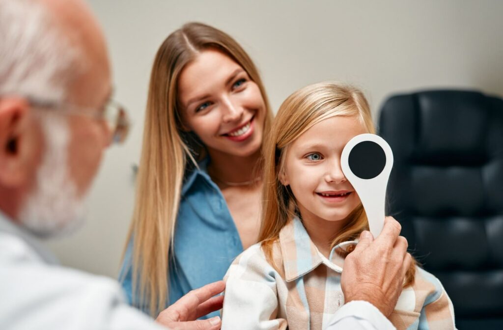 eye doctor covering smiling little girl's left eye during eye exam while her mother happily supervises behind daughter